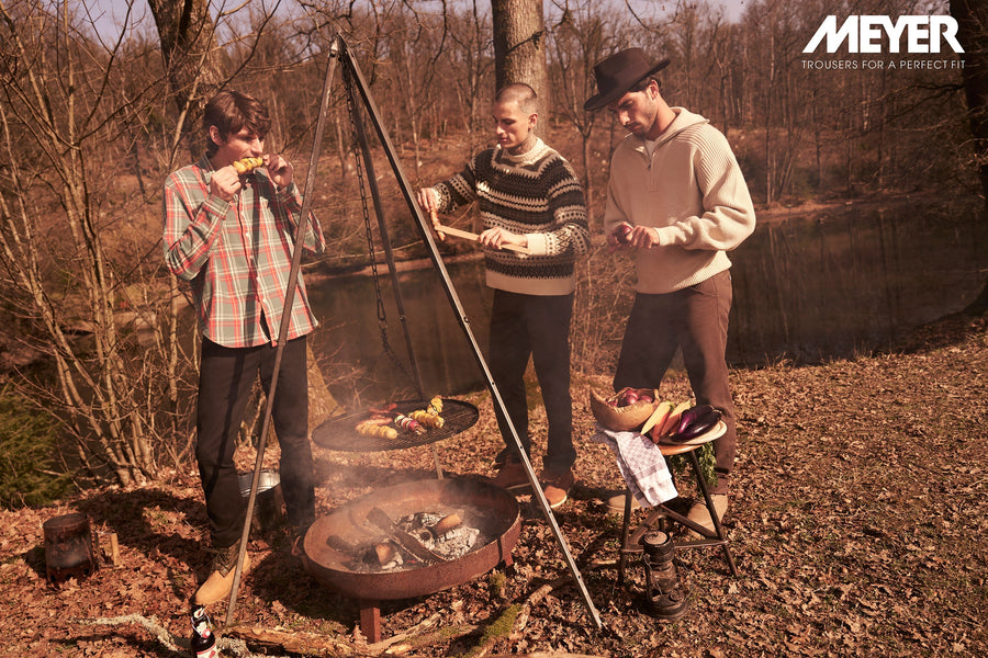Meyer campaign image showing three men outdoors by a fire wearing Meyer trousers and knitwear